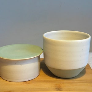 May include: A set of two ceramic butter dishes with lids. The dishes are white with a green glaze on the top and bottom. The dishes are on a wooden chopping board.