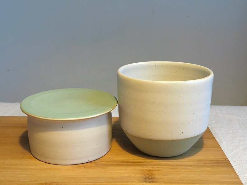 May include: A set of two ceramic butter dishes with lids. The dishes are white with a green glaze on the top and bottom. The dishes are on a wooden chopping board.