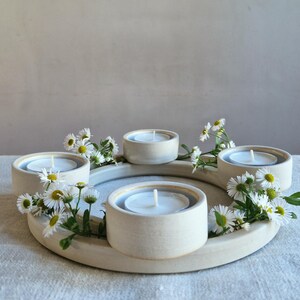 May include: A cream-colored ceramic candle holder with five circular wells, each holding a lit white tea light candle. The holder is decorated with small white and yellow daisies. The arrangement is on a light-colored surface.
