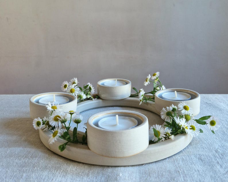 May include: A cream-colored ceramic candle holder with five circular wells, each holding a lit white tea light candle. The holder is decorated with small white and yellow daisies. The arrangement is on a light-colored surface.