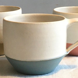 May include: Three ceramic mugs with different colored bands around the bottom. The mugs are white with a light blue, yellow, and pink band.