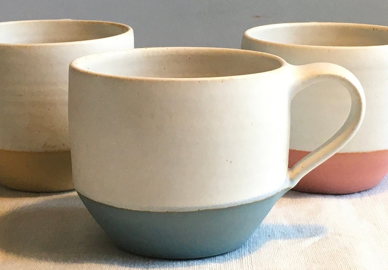 May include: Three ceramic mugs with different colored bands around the bottom. The mugs are white with a light blue, yellow, and pink band.