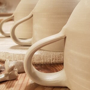 May include: Three unglazed ceramic mugs with handles, sitting on a white round surface. The mugs are in various stages of completion, with one mug having a handle that is still wet and unfinished.