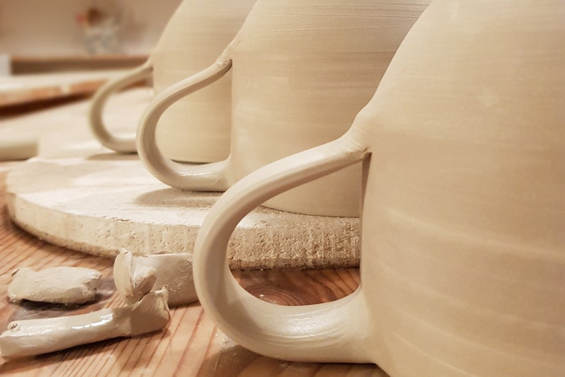 May include: Three unglazed ceramic mugs with handles, sitting on a white round surface. The mugs are in various stages of completion, with one mug having a handle that is still wet and unfinished.