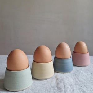 Könnte beinhalten: Vier Eierbecher aus Keramik in einer Reihe, die jeweils ein braunes Ei halten. Die Becher haben eine matte Oberfläche und sind in Hellgrün, Creme, Blau und zartem Rosa gehalten. Jeder Becher hat eine konische Form mit einem flachen Boden.