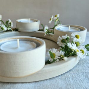 May include: A cream-colored ceramic candle holder with three circular wells, each holding a white tea light candle. White daisy flowers with yellow centers are arranged around the holder, creating a natural, decorative display.