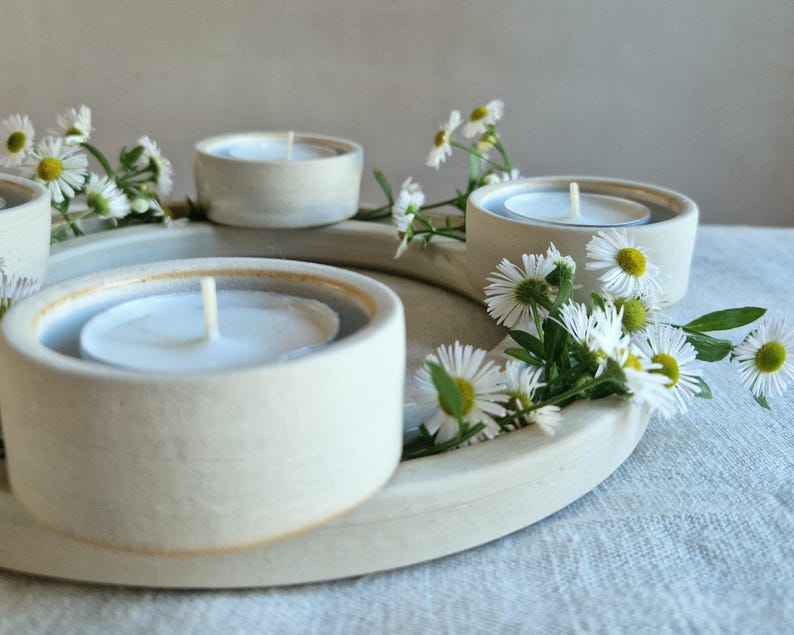 May include: A cream-colored ceramic candle holder with three circular wells, each holding a white tea light candle. White daisy flowers with yellow centers are arranged around the holder, creating a natural, decorative display.