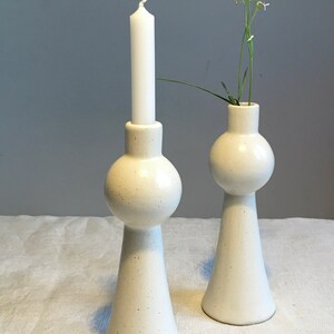 Op de afbeelding: Twee witte keramische kaarsenstandaards met een gespikkelde afwerking. De kaarsenstandaard links heeft een witte kaars. De kaarsenstandaard rechts heeft een klein boeket witte bloemen.