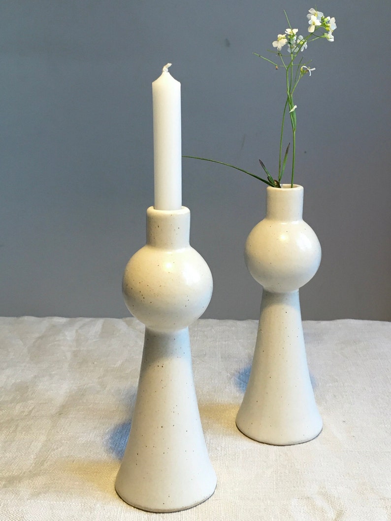 Op de afbeelding: Twee witte keramische kaarsenstandaards met een gespikkelde afwerking. De kaarsenstandaard links heeft een witte kaars. De kaarsenstandaard rechts heeft een klein boeket witte bloemen.