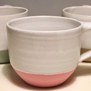 May include: Three ceramic mugs with different colored glazes. The mug on the left is light green, the mug in the center is pink, and the mug on the right is yellow.