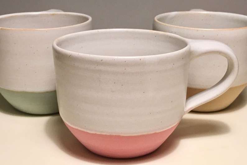 May include: Three ceramic mugs with different colored glazes. The mug on the left is light green, the mug in the center is pink, and the mug on the right is yellow.