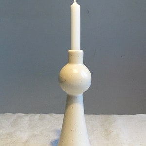 Op de afbeelding: Witte keramische kaarsenhouder met een ronde, bolle bovenkant en een uitlopende basis. De houder is bezaaid met kleine bruine vlekjes.