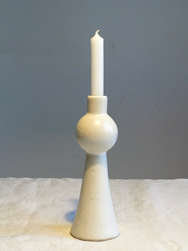 Op de afbeelding: Witte keramische kaarsenhouder met een ronde, bolle bovenkant en een uitlopende basis. De houder is bezaaid met kleine bruine vlekjes.