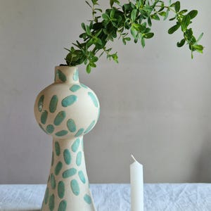 Op de afbeelding: Een witte keramische vaas met een groen abstract patroon, gevuld met groene bladeren. Een witte kaars staat op een gouden kaarsenhouder.