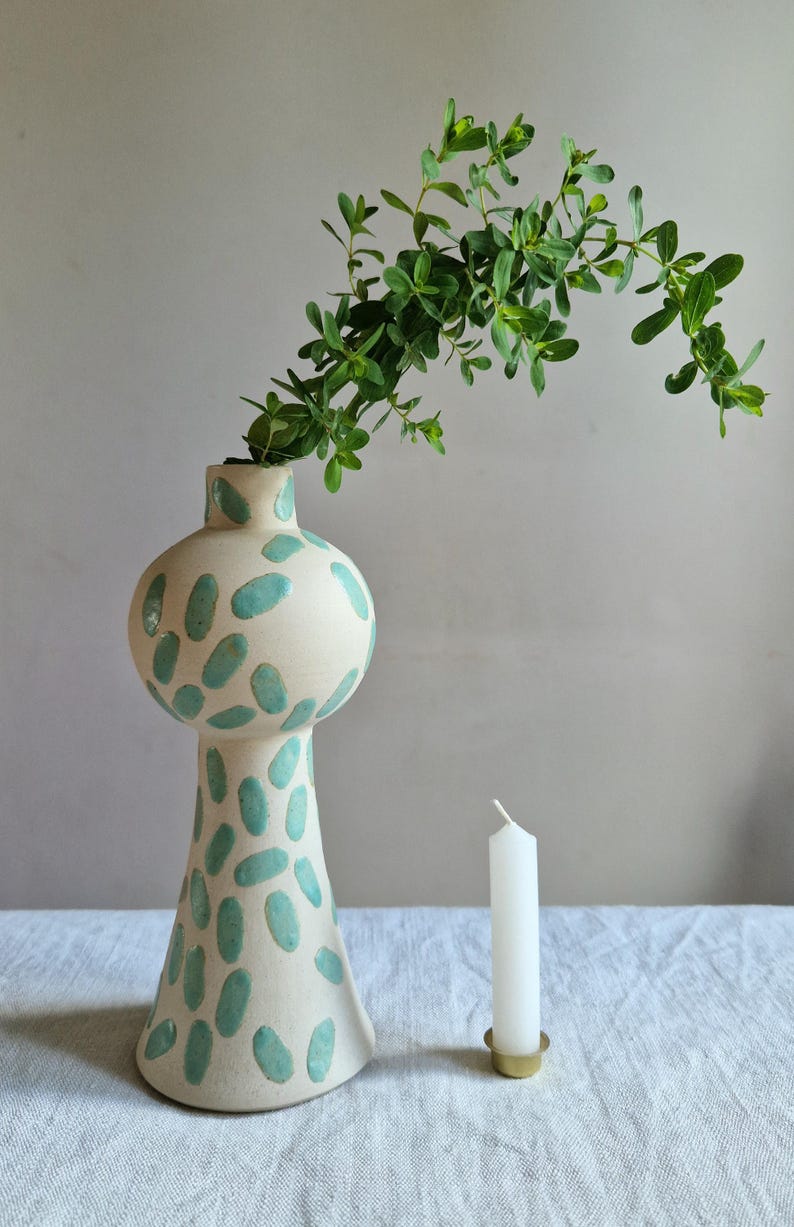 Op de afbeelding: Een witte keramische vaas met een groen abstract patroon, gevuld met groene bladeren. Een witte kaars staat op een gouden kaarsenhouder.