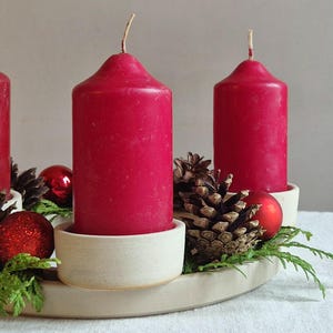 May include: A festive Advent wreath with four red candles. The candles sit in off-white holders, surrounded by pine cones, red ornaments, and greenery. The wreath is on a white surface.