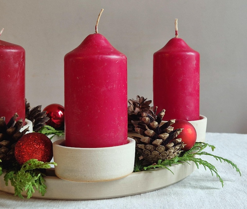 May include: A festive Advent wreath with four red candles. The candles sit in off-white holders, surrounded by pine cones, red ornaments, and greenery. The wreath is on a white surface.