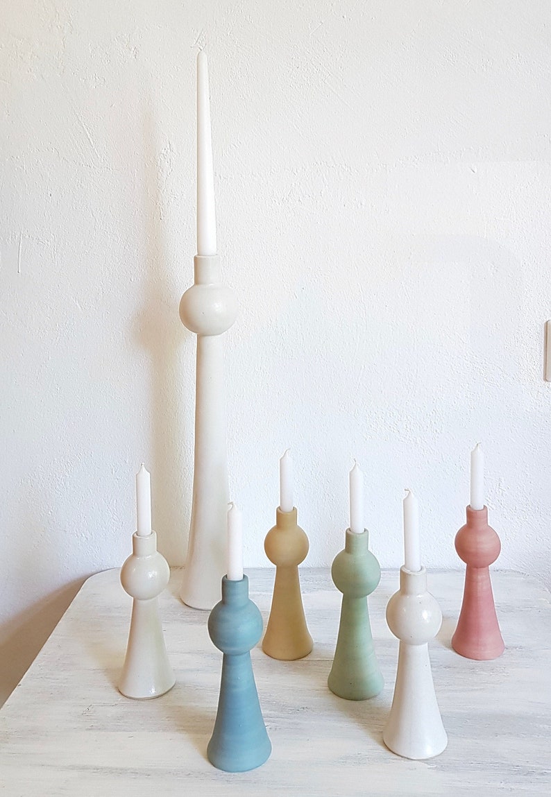 Op de afbeelding: Set van zeven keramische kaarsenstandaards in verschillende pastelkleuren, waaronder wit, lichtblauw, groen, geel en roze. De standaards hebben allemaal de vorm van een hoge, slanke vaas met een ronde bol aan de bovenkant.