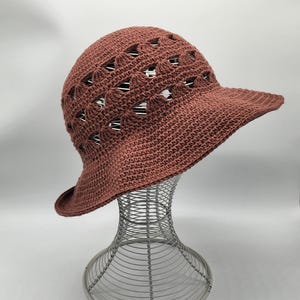 May include: A brown crocheted sun hat with a wide brim and a lacy design. The hat is on a wire hat stand.