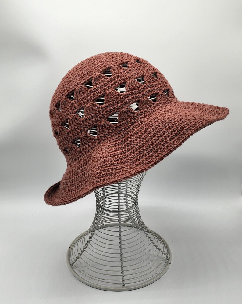 May include: A brown crocheted sun hat with a wide brim and a lacy design. The hat is on a wire hat stand.