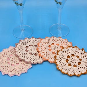 May include: A collection of five handmade crochet coasters in various pastel colors, including pink, cream, and peach, arranged on a blue surface. The coasters feature a detailed floral design and are placed near two clear champagne flutes.