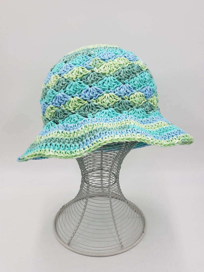 May include: A crocheted sun hat with a wide brim in shades of green and blue. The hat is displayed on a wire hat stand.