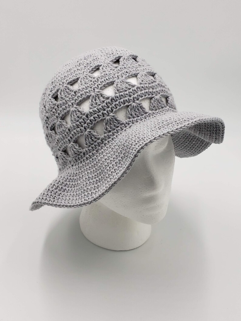 May include: A grey crochet sun hat with a wide brim and a geometric pattern. The hat has openwork details, creating a lightweight and airy design.