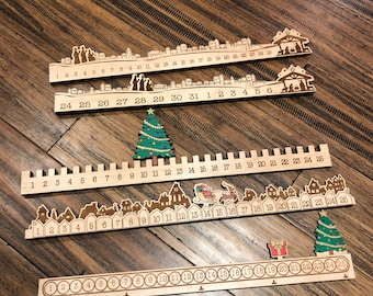 Può includere: Quattro calendari dell'avvento in legno con i numeri da 1 a 25. Ogni calendario presenta una scena natalizia diversa con case, un albero di Natale e una scena della natività.
