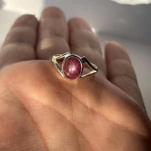 May include: A silver ring with an oval ruby gemstone displaying a star effect. The ring's band has an open design with geometric cutouts. The ruby is a deep red colour with a star pattern. The ring is a piece of jewellery.