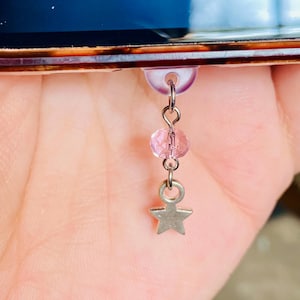 Pode incluir: Um pequeno pingente de estrela prateada pende de uma corrente, conectado a uma conta rosa e uma conta roxa. O pingente está preso a um objeto marrom e preto.