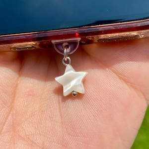 Può includere: Un ciondolo a forma di stella bianca perlescente con una perla color argento sul fondo. La stella è attaccata a una custodia trasparente per telefono con un attacco viola. Lo sfondo è una mano sfocata ed erba verde.