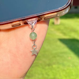 May include: A silver phone charm with a green bead and a star-shaped charm. The charm is attached to a clear phone case. The background is blurred, showing green grass.