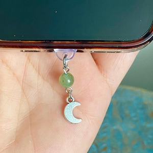 May include: A phone charm featuring a small, silver crescent moon pendant and a green gemstone bead. The charm is attached to a clear suction cup, designed to adhere to a phone. The charm is suspended from a silver chain.