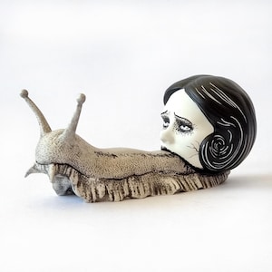 Puede incluir: Una escultura de cerámica blanca de un caracol con una cabeza humana. La cabeza está pintada con cabello negro y una cara blanca con ojos negros y una boca negra. El caracol se arrastra sobre una superficie blanca.