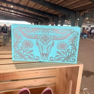 May include: A turquoise rectangular box with a silver steer skull and floral design. The box is displayed on a wooden crate, with a pair of pink and brown shoes in the foreground. The background shows a large indoor space.
