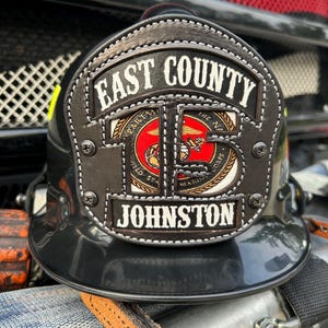 May include: A black firefighter helmet with a leather front shield. The shield has the words "EAST COUNTY" and "JOHNSTON" in white lettering. A circular emblem with a red background and gold accents is centered on the shield.
