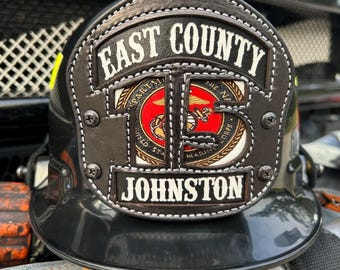May include: A black firefighter helmet with a leather front shield. The shield has the words "EAST COUNTY" and "JOHNSTON" in white lettering. A circular emblem with a red background and gold accents is centered on the shield.