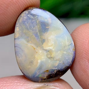 May include: A polished opal gemstone with a teardrop shape. The stone features a mix of colors, including white, blue, yellow, and brown. The opal is held between two fingers, showcasing its iridescent surface and natural patterns.