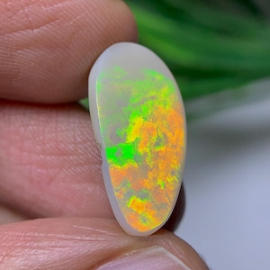 Può includere: Una gemma di opale bianco a forma di goccia caratterizzata da un vibrante gioco di colori con luminosi bagliori verde neon e arancione acceso su tutta la superficie. La pietra presenta una finitura liscia e un taglio cabochon rotondo.