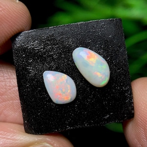 May include: Two pear-shaped opals with a milky white base and flashes of orange, yellow, and green. The opals are set against a dark, textured surface, showcasing their iridescent qualities. These gemstones are ideal for jewelry making.