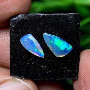 May include: Two polished opal gemstones with vibrant blue, green, and yellow hues. The opals are set against a black background, showcasing their iridescent play of color. The gemstones have a teardrop shape.