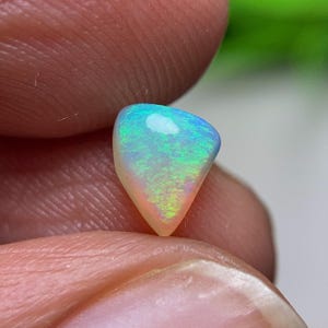 Puede incluir: Una piedra preciosa de ópalo triangular pulida. El ópalo muestra un vibrante juego de colores, con tonos azules, verdes y amarillos y un brillo nacarado. La piedra preciosa se sostiene entre los dedos, mostrando su belleza natural.