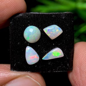May include: Four opals of varying shapes and sizes are displayed on a black surface. The opals exhibit a play of color, with flashes of green, blue, and red. One is round, one is a diamond shape, and the other two are triangular.