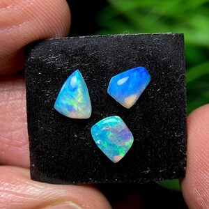 Könnte beinhalten: Drei polierte Opal-Edelsteine in Blau-, Grün- und Weißtönen. Die Opale sind auf einer schwarzen Oberfläche angeordnet. Die Edelsteine haben einen perlmuttartigen Glanz und sind unregelmäßig geformt.