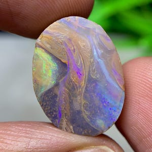 May include: An oval opal gemstone with a swirling pattern of colours. The opal displays hues of purple, blue, green, and tan. The stone is held between two fingers, showcasing its natural, raw edges and unique colour play.