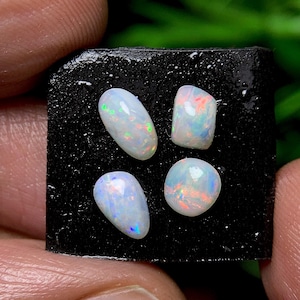May include: Four opals with iridescent color flashes are displayed on a black surface. The opals vary in shape, including oval and rounded forms. The opals exhibit a play of color, with hues of green, blue, and red.
