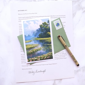 May include: A landscape art print titled "Aquatic Reflection" showing a tranquil lake scene with trees and flowers. The print is on a white card, with a green envelope, a pen, and a letter. The letter is signed and dated September 2025.