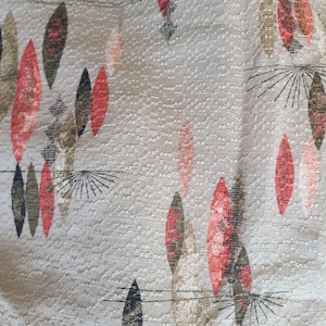May include: Close-up of a textured fabric featuring a repeating pattern of stylized leaf shapes in red, black, and olive green. The leaves are set against a light beige background, with thin black lines.