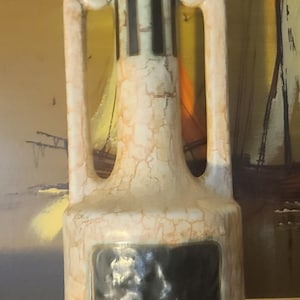 May include: A cream-colored ceramic vase with a cracked glaze effect. The vase has a rectangular black panel on the front and two curved handles. The top features vertical black stripes.