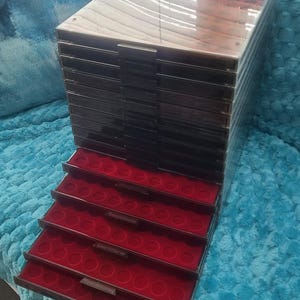 May include: A black coin storage cabinet with a clear top and multiple stacked drawers. The interior of the open drawers features a vibrant red velvet lining with circular indentations designed to hold individual coins securely in place.